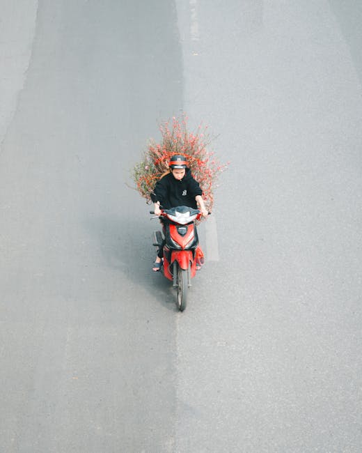 A person wearing a black helmet and dark clothing is riding a red motorbike on a paved surface. The motorbike is carrying a large bouquet of mixed flowers with pink, orange, and green hues, arranged w