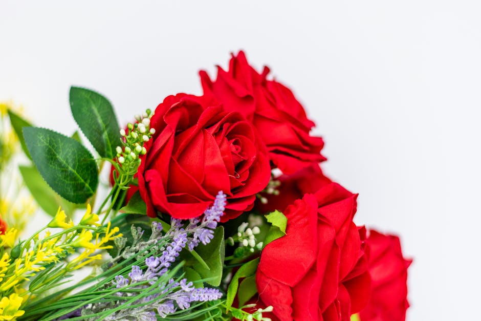 A bouquet featuring vibrant red roses with multiple layered petals, complemented by green foliage with broad, textured leaves. Additionally, small sprigs of purple lavender and tiny white flowers add