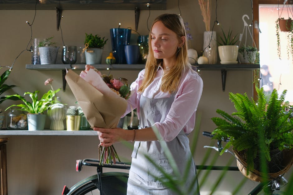 A young woman with long, light brown hair is standing in a cozy, well-lit room decorated with fairy lights, holding a floral bouquet wrapped in brown paper. She is dressed in a light pink blouse and a