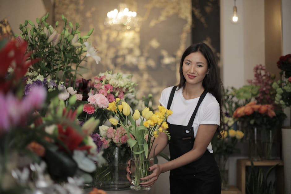 A young woman with long dark hair, dressed in a white t-shirt and black apron, is standing inside a flower shop with the logo 'flowersharrow.com'. She is smiling gently while holding a glass vase fill