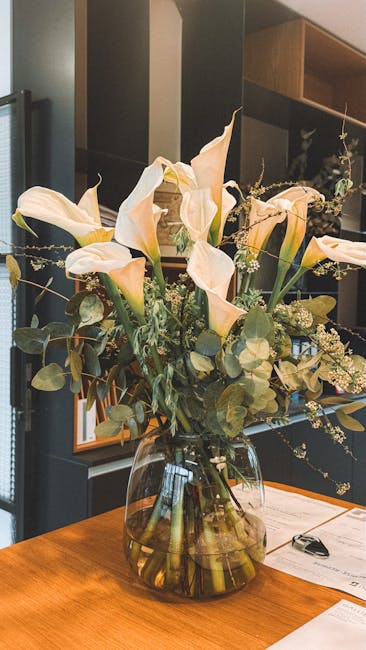 A tall, elegant bouquet of fresh cream-colored calla lilies with smooth, trumpet-shaped petals and long, slender stems, complemented by sprigs of small white filler flowers and round, silvery-green eu