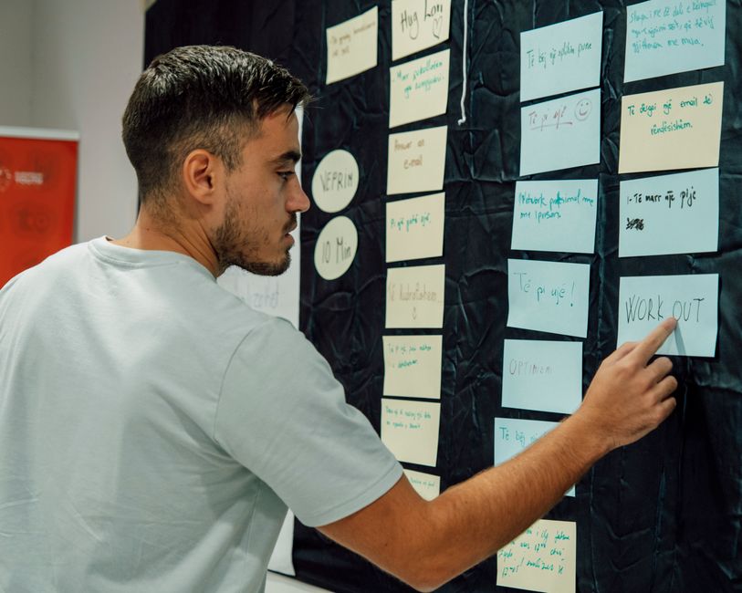 A young man with short, dark hair and a light-colored t-shirt is placing a light blue note on a black wall covered with various notes and sticky notes. The notes are arranged in columns and contain ha