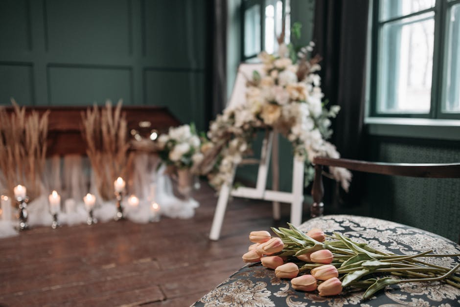 A bouquet of soft pink tulips with green stems and leaves rests on an ornate, dark-colored upholstered chair in a softly lit room. In the background, a funeral floral arrangement with white flowers an