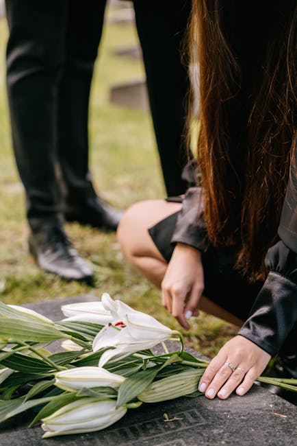 A close-up of a woman in black clothing kneeling on the ground, gently placing her hand on a large bouquet of white lilies with green leaves, which are laid on a dark stone surface during a somber out