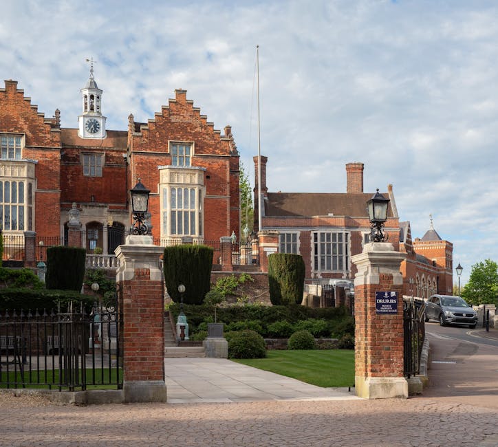 The image shows a historic red-brick mansion with ornate architectural details, large sash windows, and a prominent clock tower topped with a weather vane, set against a partly cloudy sky. The mansion