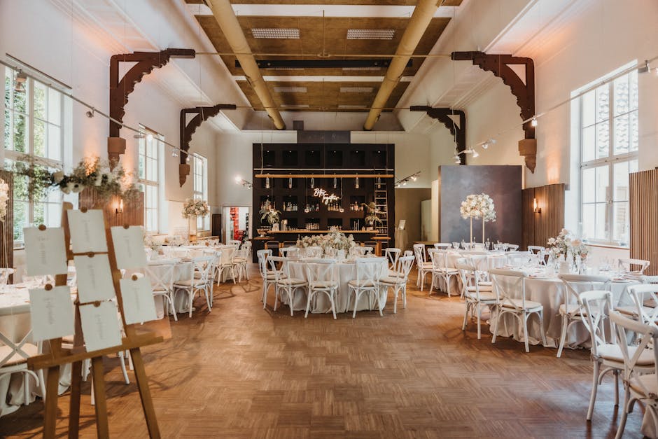 A spacious interior decorated for a wedding reception at Harrow School venue, featuring several round tables covered with elegant white tablecloths and adorned with floral centerpieces consisting of w